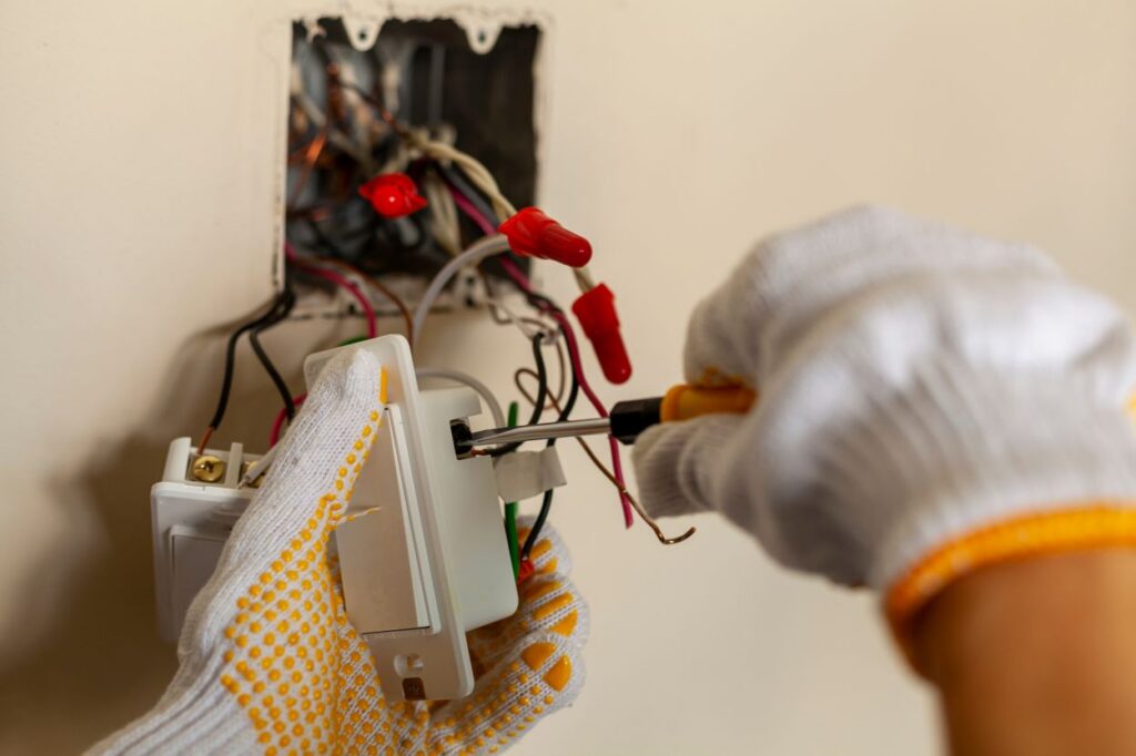An electrician is replacing a wall switch. A DIY project concept. High voltage danger. installing wire connection using screw driver. The professional wears protective rubber gloves for safety See Less